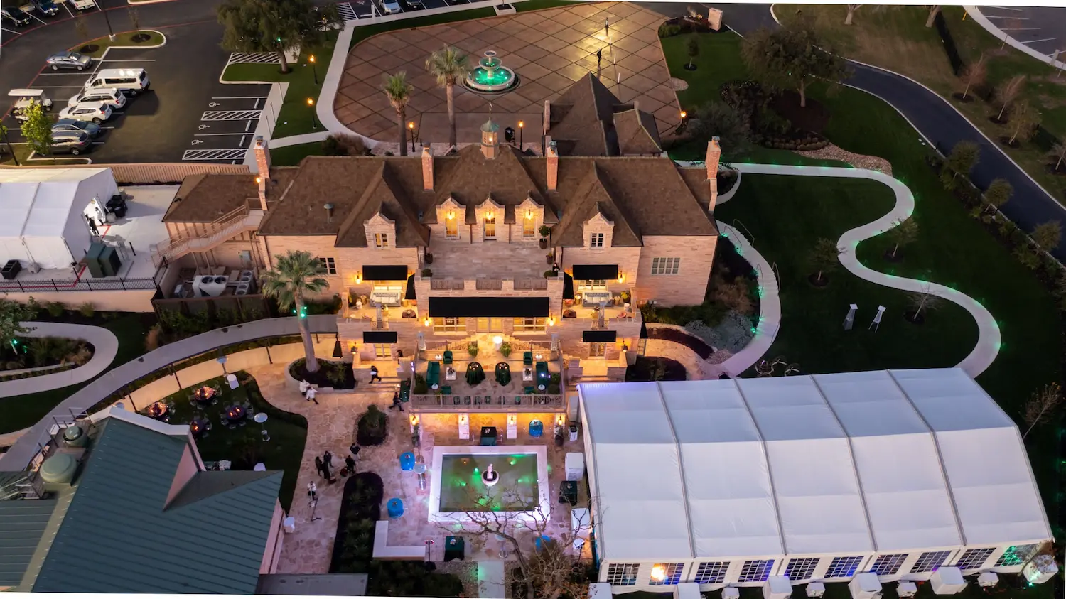 Aerial view of The Red Berry Estate mansion at dusk, showcasing illuminated outdoor terraces, fountains, and landscaped grounds.