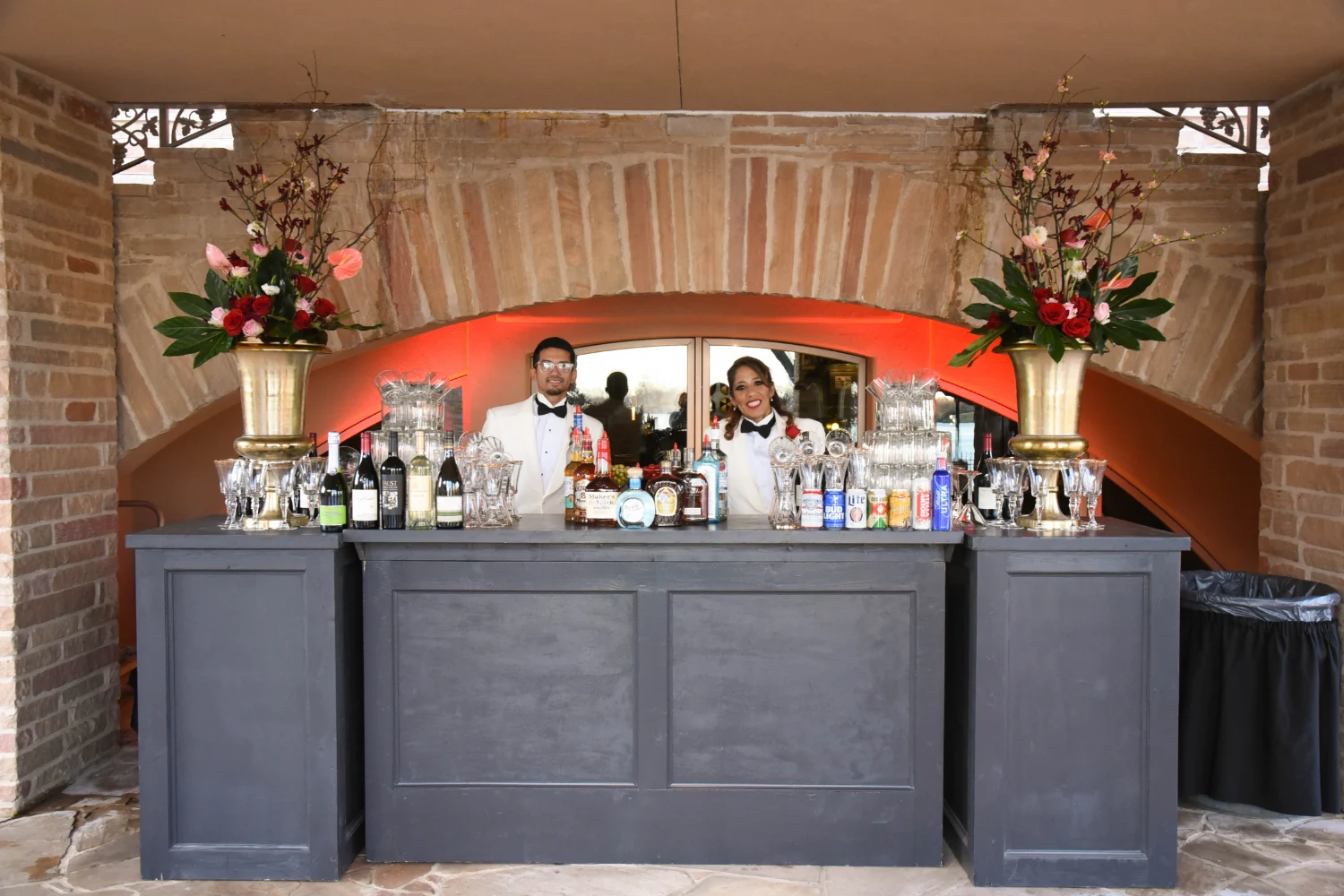 Two bartenders smiling at the bar with a variety of liquors and beers