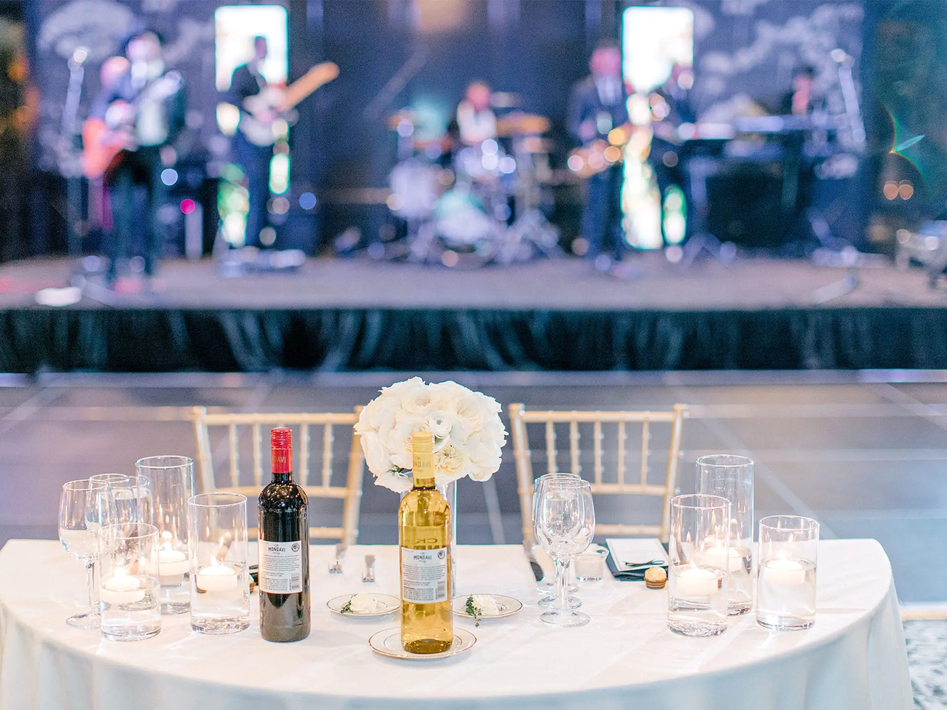 Elegant sweetheart table set with wine, candles, and white floral arrangement in front of a live band performance at the Lakeside Ballroom at The Red Berry Estate wedding reception.
Photo: Allison Jeffers Photography