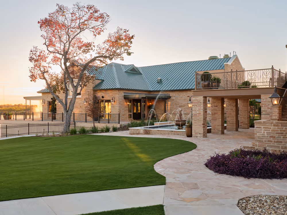 Modern lakeside pavilion with green metal roof, stone exterior, outdoor fountain, and landscaped lawn at sunset.