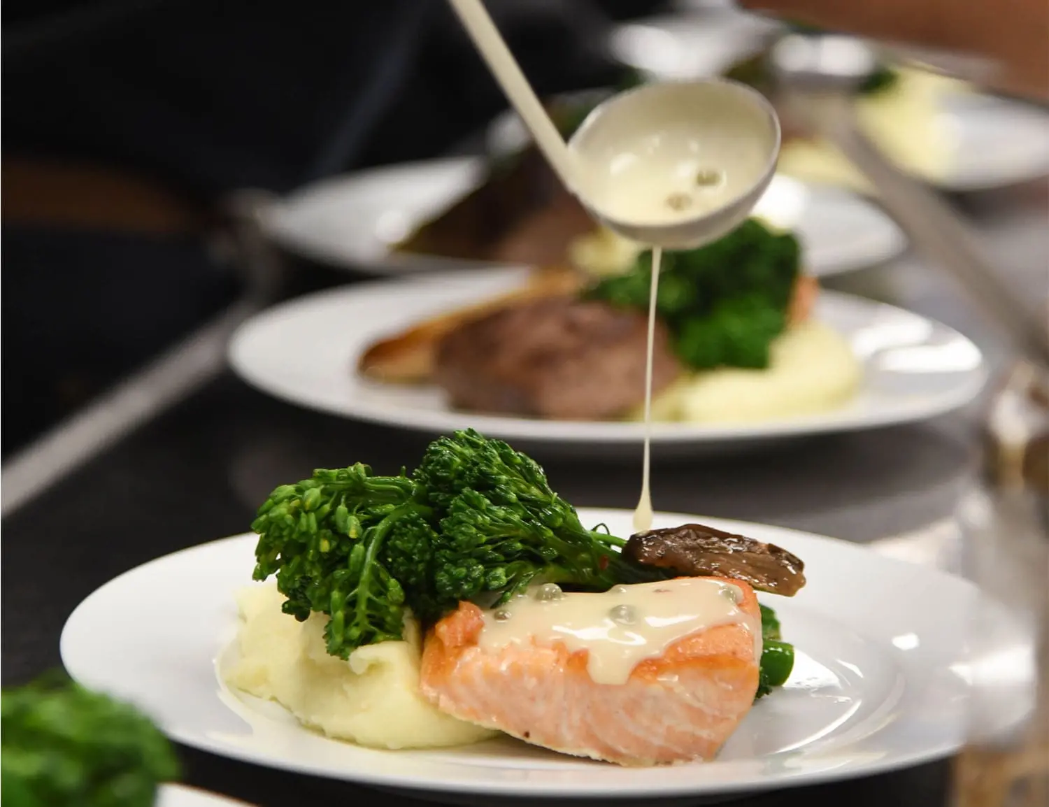 A close-up of a chef pouring creamy sauce over a plate of poached salmon, mashed potatoes, and broccolini, creating an elegant, gourmet presentation.