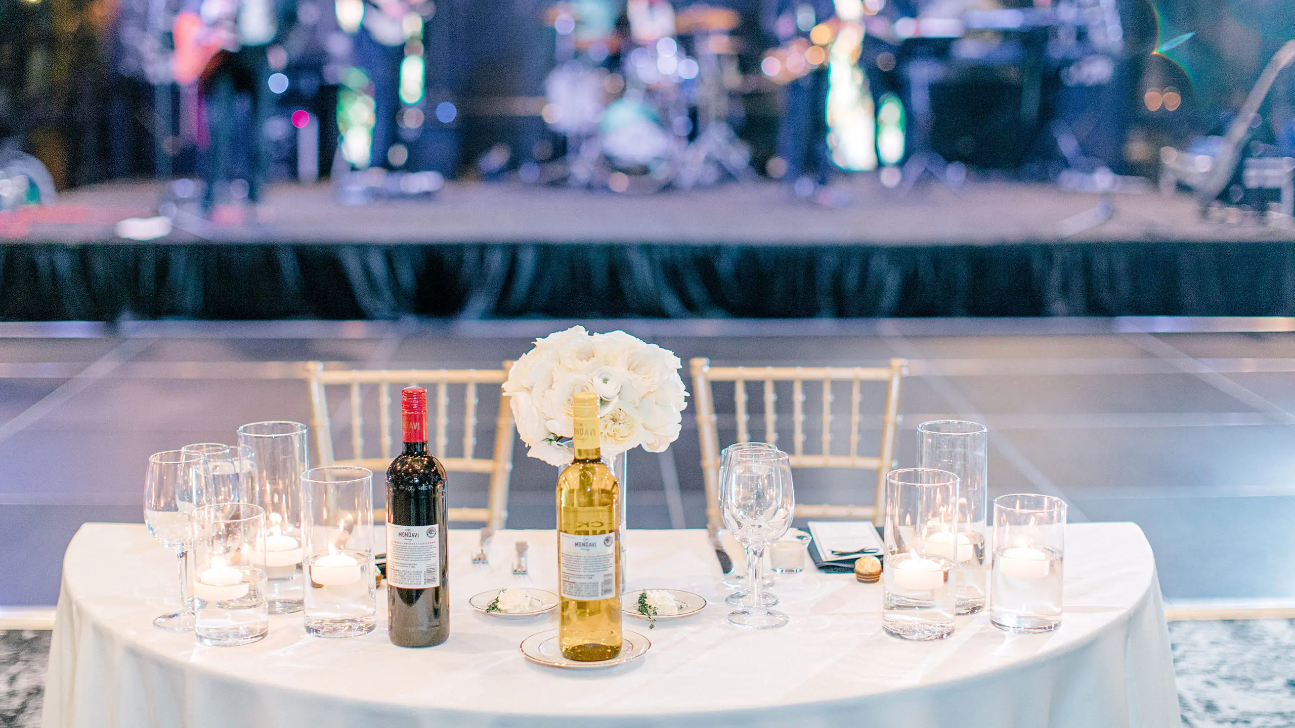 Elegant sweetheart table set with wine, candles, and white floral arrangement in front of a live band performance at the Lakeside Ballroom at The Red Berry Estate wedding reception.
Photo: Allison Jeffers Photography