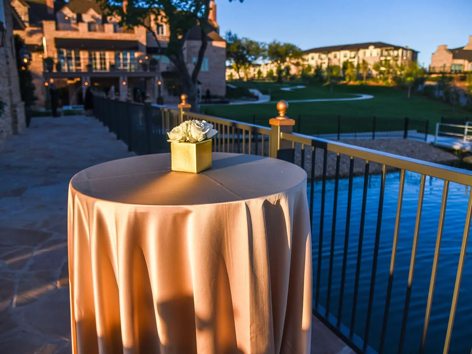 Elegant outdoor patio scene at sunset; a round table with a beige cloth and white roses. Stone path leads to a grand manor and scenic pond.