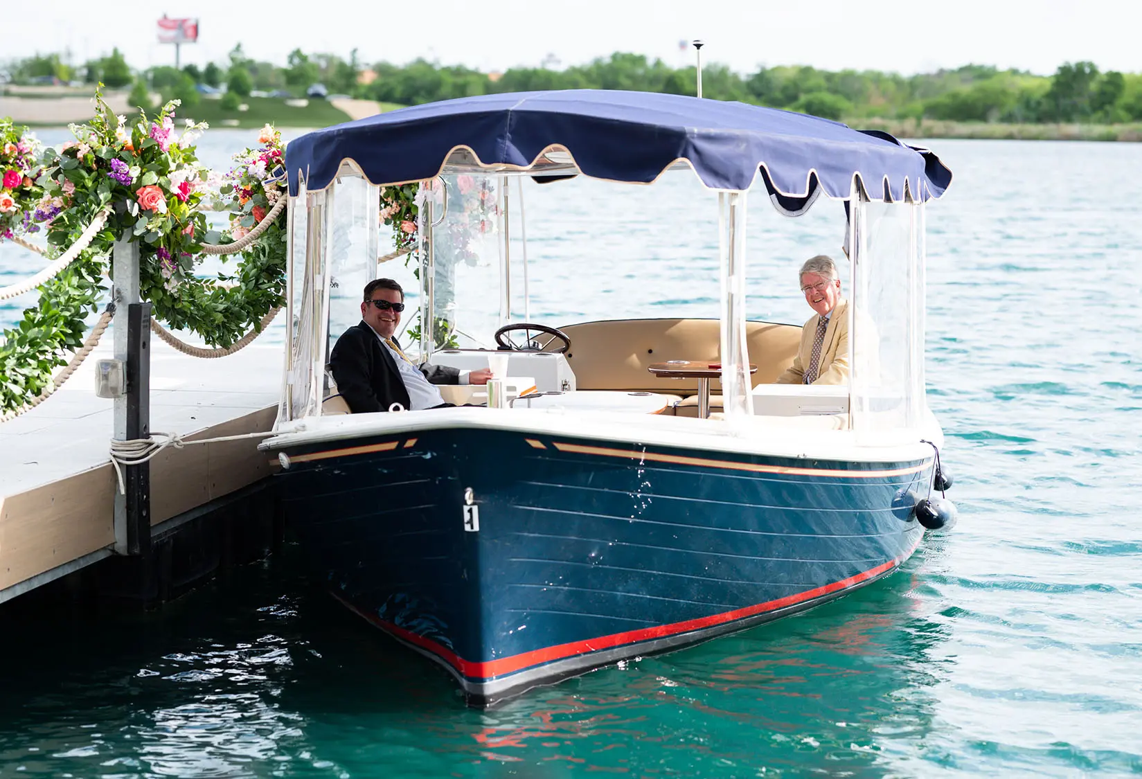 Elegant navy electric boat docked lakeside with guests smiling and seated, surrounded by floral garlands at The Red Berry Estate.
Photo: Parish Photography