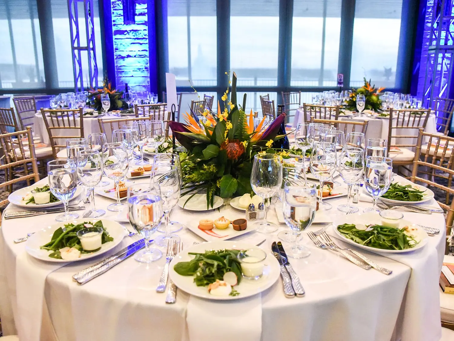 Elegant banquet table set with crisp white linens, lush floral centerpiece, and neatly arranged salads. Ambient blue lighting creates a sophisticated atmosphere. The Red Berry Estate in San Antonio, Texas.