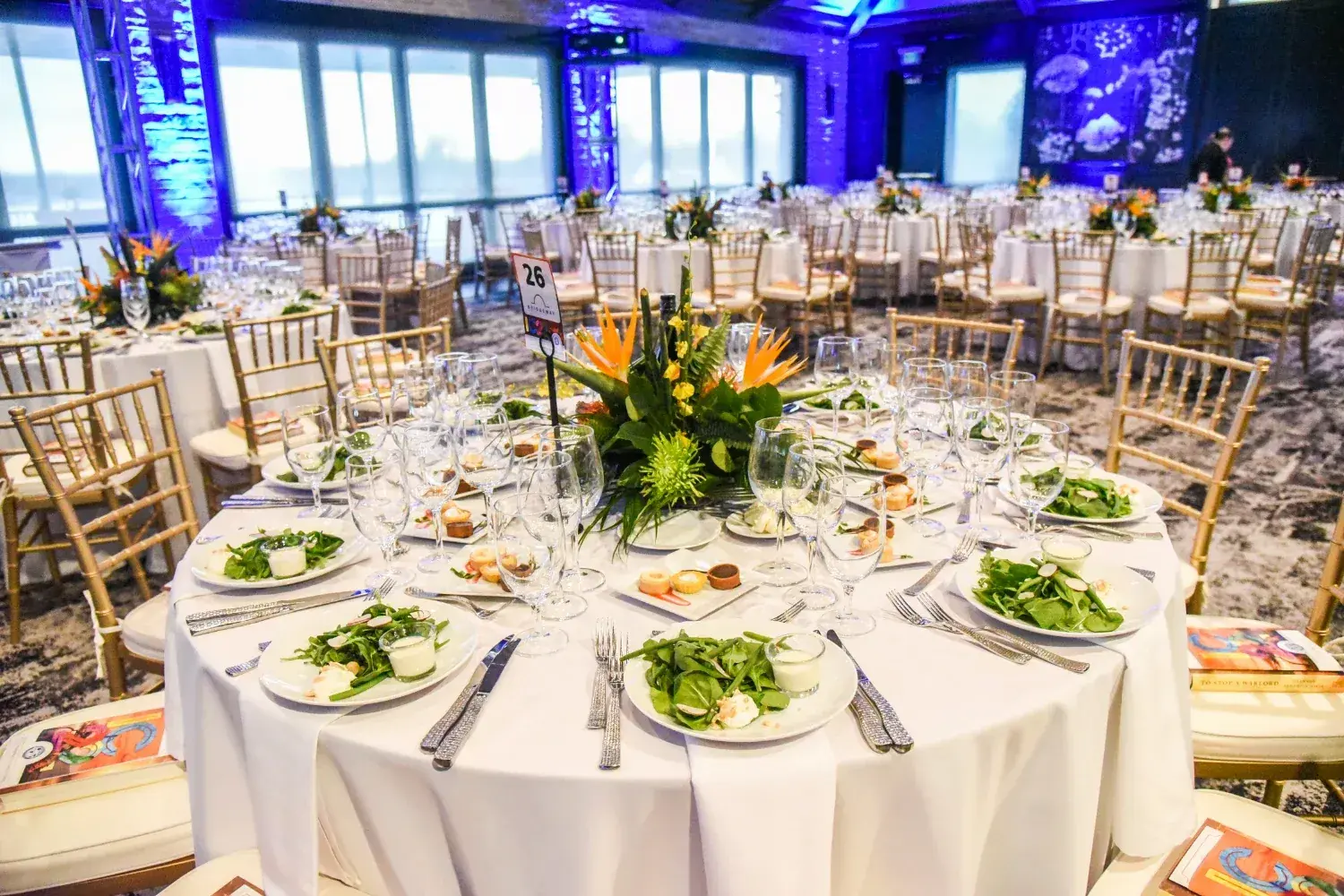 Round tables set for a formal nonprofit gala with plated salads, floral centerpieces, and gold chairs at The Red Berry Estate.